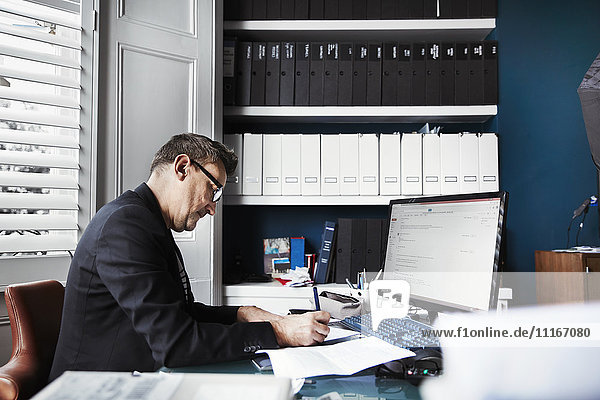 Ein Mann sitzt an einem Schreibtisch in einem Büro und schreibt auf einem Blatt Papier. Ordentliche Aktenreihen in den Regalen und Papiere auf dem Schreibtisch.