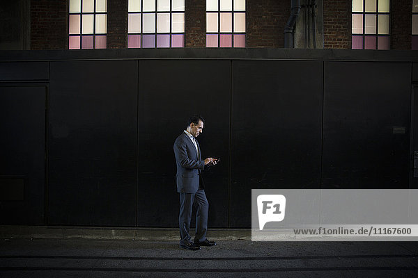 Ein Mann in einem Anzug steht im Schatten auf einer Straße der Stadt unter einem erleuchteten Fenster und überprüft sein Telefon.