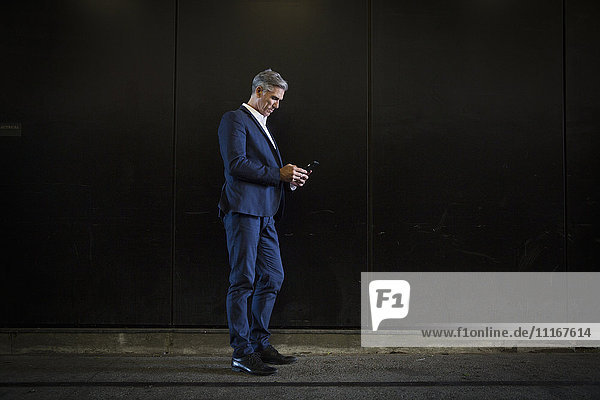Ein Mann in einem Anzug  der im Schatten auf einer Straße der Stadt steht  auf sein Smartphone schaut  eine SMS schreibt oder online nach Informationen sucht.