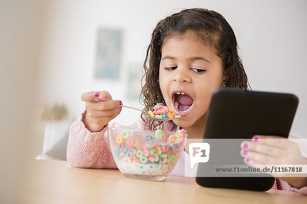 Gemischtrassiges Mädchen isst Müsli und benutzt ein digitales Tablet