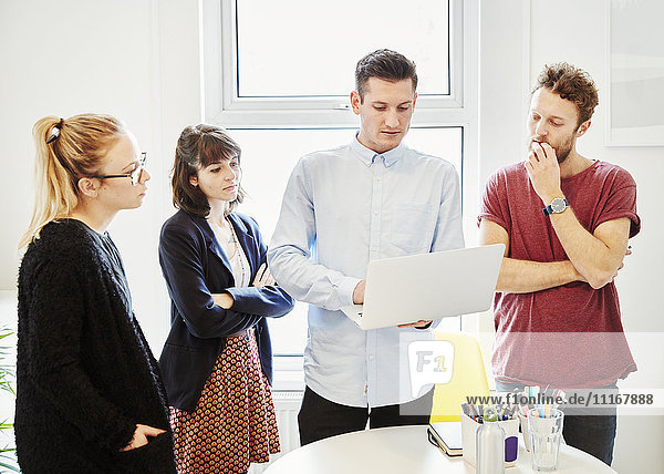 Vier Personen stehen bei einer Geschäftsbesprechung um einen Tisch herum und schauen auf einen Laptop-Bildschirm.
