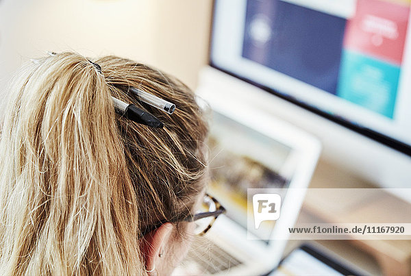 Über-die-Schulter-Ansicht einer Frau an einem Arbeitsplatz mit zwei Stiften im Haar. Laptop- und Computerbildschirmgrafiken.