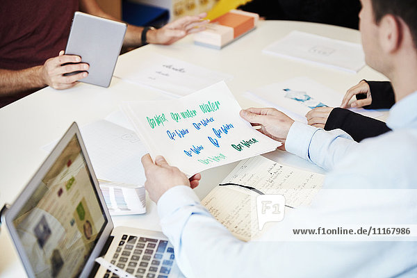 Ein Mann hält Papier mit gedruckten Logos und Schriftzügen in verschiedenen Stilen und Farben in der Hand. Notizbücher  ein Laptop und ein digitales Tablett. Designer bei der Arbeit.