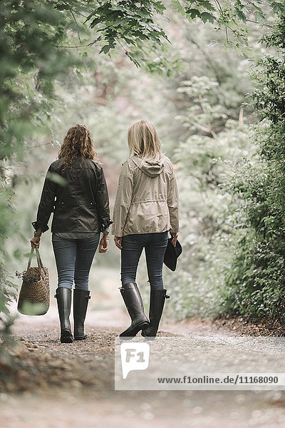 Zwei Frauen gehen in Mänteln und Stiefeln mit einem Korb einen Feldweg entlang.