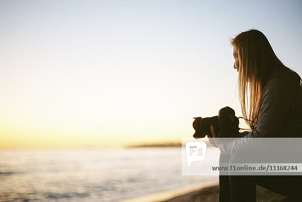 Seitenansicht einer Frau mit langen blonden Haaren  die an einem Sandstrand sitzt und eine Kamera hält.