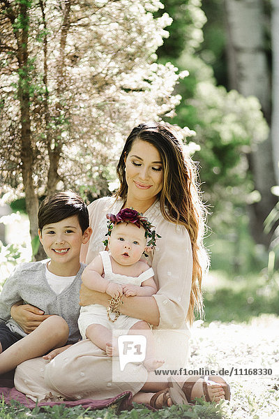 Porträt einer lächelnden Mutter  eines Jungen und eines kleinen Mädchens mit einem Blumenkranz auf dem Kopf  die in einem Garten sitzen.