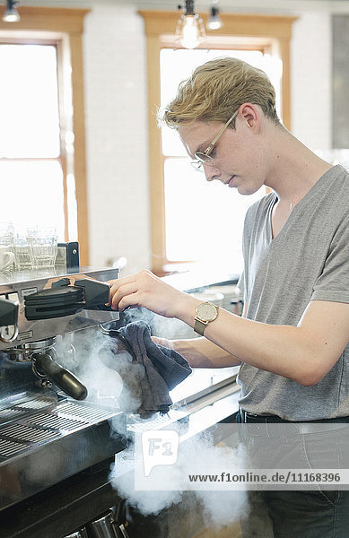 Junger blonder Mann mit Brillengestell steht vor einer Espressomaschine  aus der Dampf austritt.