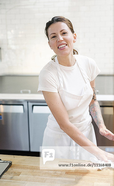 Frau mit weißer Schürze steht an einem Arbeitstresen in einer Bäckerei und wischt die Oberfläche mit einem Tuch ab.