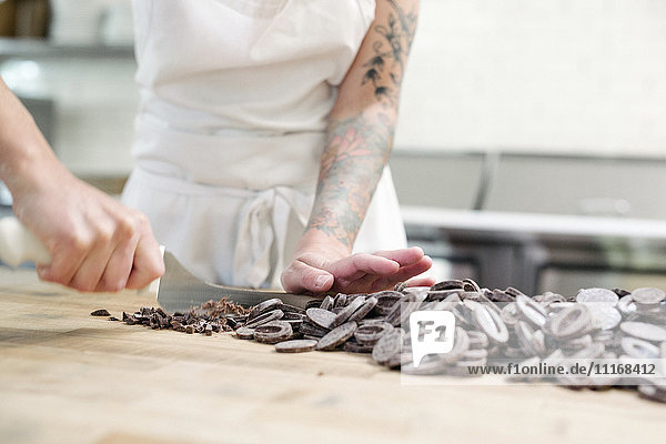 Nahaufnahme einer Frau mit einer weißen Schürze  die an einem Arbeitstresen in einer Bäckerei steht und Schokolade schneidet.