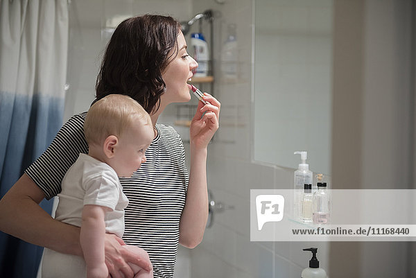 Caucasian mother holding baby son while applying lipstick in mirror
