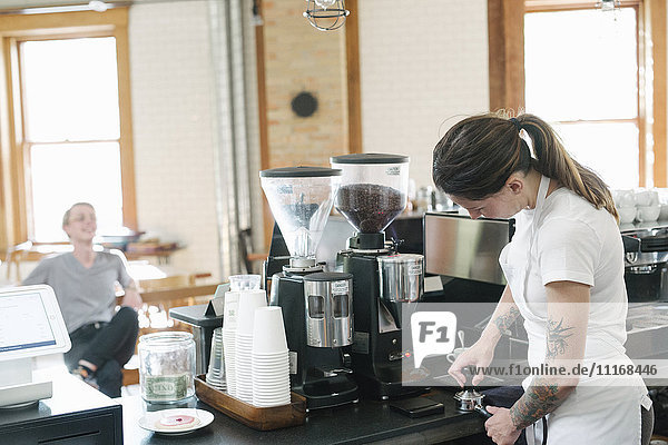 Frau mit einer weißen Schürze  die vor einer Espressomaschine steht und einen Siebträger hält.