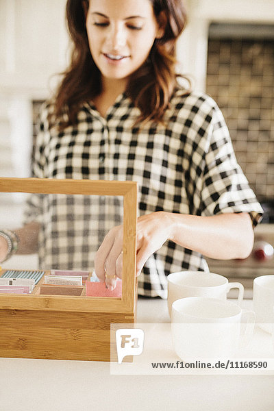 Woman with long brown hair  wearing a chequered shirt  standing in a kitchen  making a cup of tea.