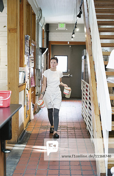 Frau mit einer Schürze  die einen Korridor entlanggeht und zwei Lebensmittelbehälter trägt.