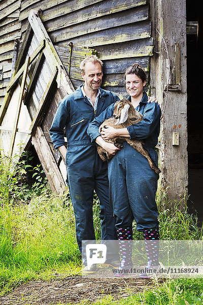 Mann und Frau stehen vor einem Stall und halten eine Ziege.