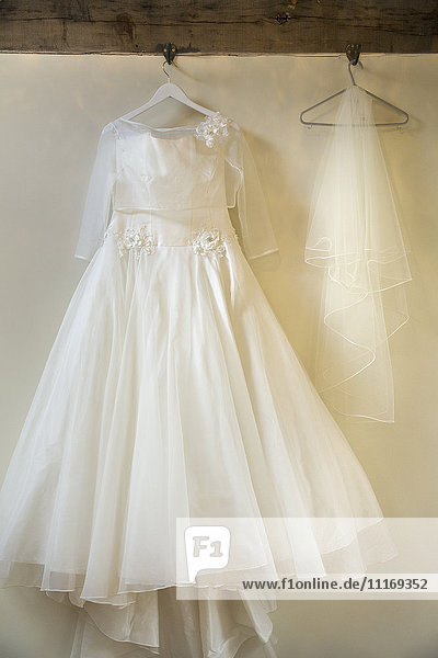 Ein langes weißes Hochzeitskleid mit vollem Rock  Petticoats und Schleier auf einem Bügel  der an einem Haken hängt.