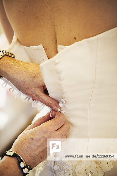 Eine Frau knöpft den Rücken des weißen Hochzeitskleides einer Braut zu.