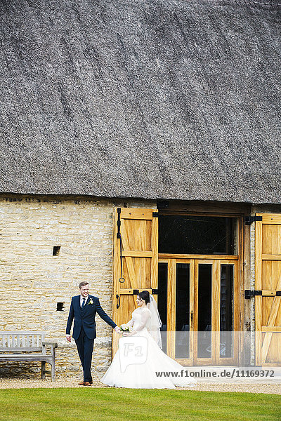Eine Braut und ein Bräutigam an ihrem Hochzeitstag  die Hand in Hand aus einer Scheune gehen.