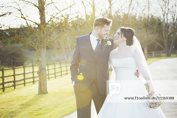 Eine Braut und ein Bräutigam  die an ihrem Hochzeitstag Arm in Arm einen Weg im Sonnenschein lachend hinuntergehen.