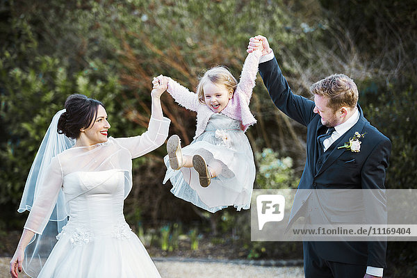 Eine Braut und ein Bräutigam  ein Paar  das an seinem Hochzeitstag ein kleines Mädchen in der Luft zwischen sich hält und schaukelt.