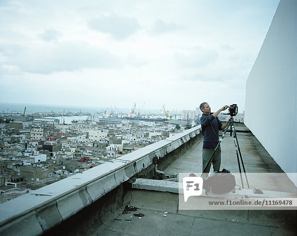 Fotograf  der auf einem Dach in einer Stadt steht und seine Kamera auf einem Stativ aufstellt.