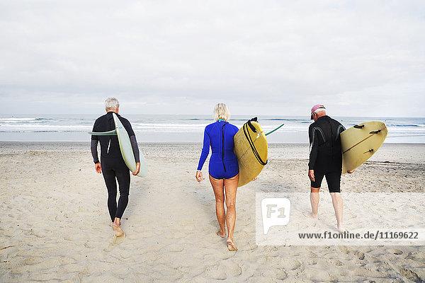 Senior woman and two senior men on a beach  wearing wetsuits and carrying surfboards.