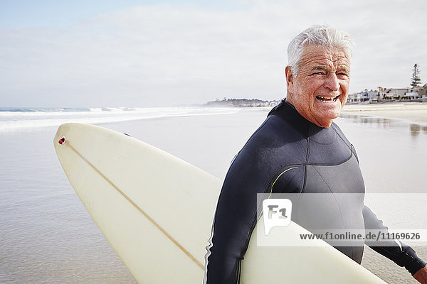 Lächelnder älterer Mann steht am Strand  trägt einen Neoprenanzug und hat ein Surfbrett dabei.