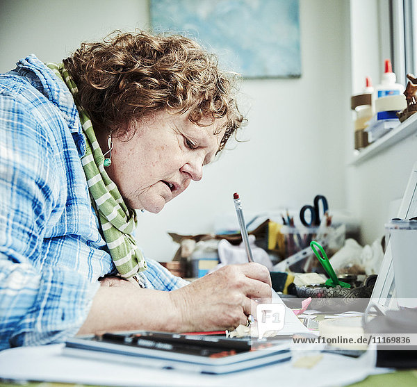 Eine Künstlerin arbeitet sitzend an einem Tisch  lehnt sich nach vorne und zeichnet mit einem Bleistift.