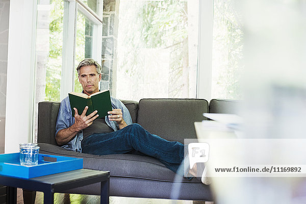 Ein Mann sitzt am Fenster auf einem Sofa und liest ein Buch.