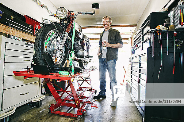 Kaukasischer Mann trinkt Kaffee  während er in der Garage ein Motorrad repariert