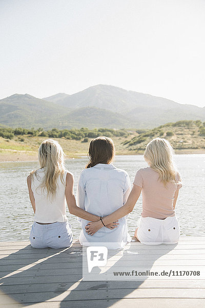 Rückansicht einer Mutter und ihrer beiden blonden Töchter  die auf einem Steg sitzen.