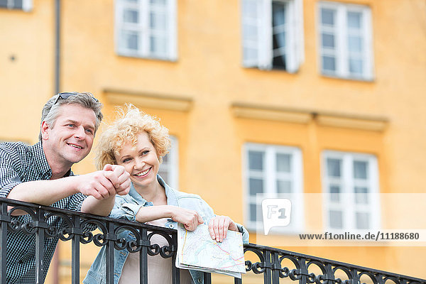Happy middle-aged couple with map leaning on railing against building