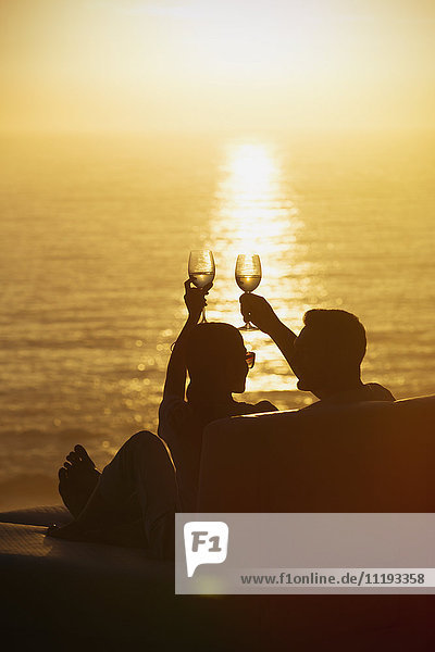 Silhouette couple toasting wine glasses on balcony with tranquil sunset ocean view