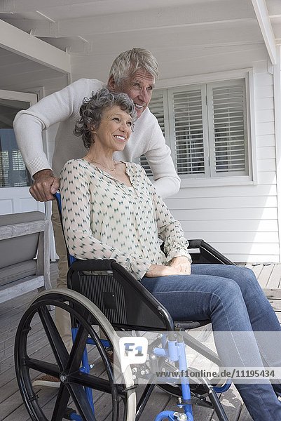 Glücklicher älterer Mann  der seiner Frau im Rollstuhl auf der Veranda hilft.