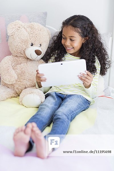 Glückliches kleines Mädchen mit einem digitalen Tablett mit Teddybär auf dem Bett