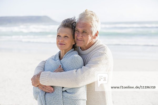 Älterer Mann  der seine Frau von hinten am Strand umarmt.