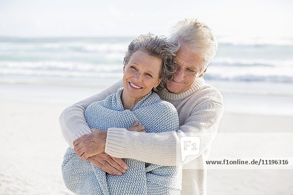 Ein älterer Mann umarmt seine Frau von hinten am Strand.