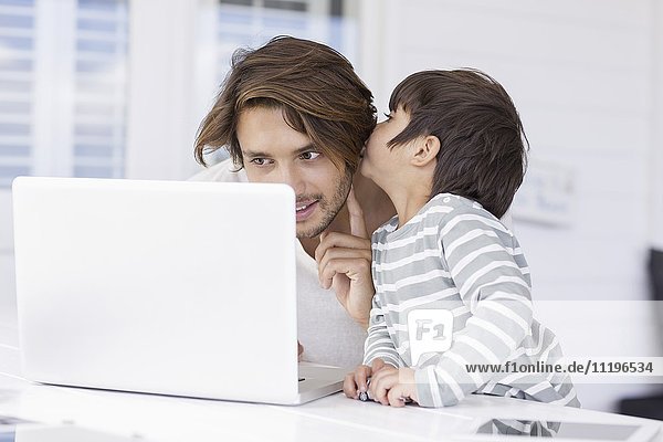 Sohn flüstert Vater ins Ohr  während er den Laptop zu Hause benutzt.