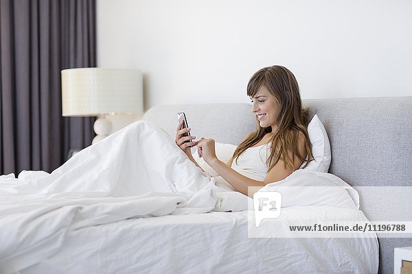 Glückliche Frau auf dem Bett sitzend mit dem Handy