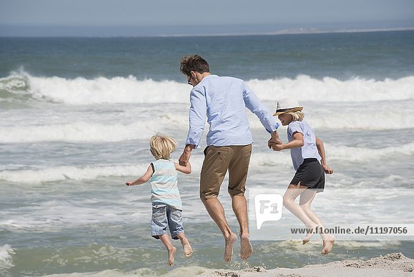 Rückansicht eines Mannes  der mit seinen Kindern am Strand sitzt.