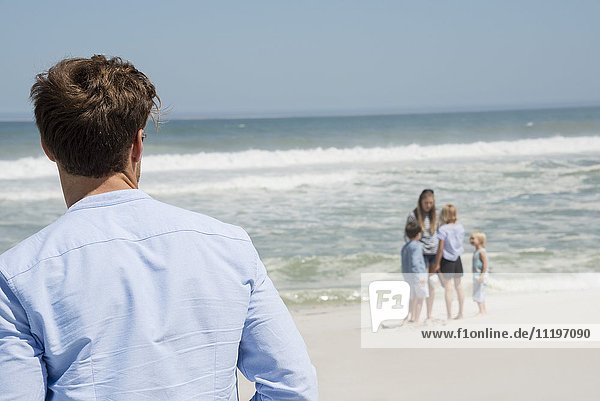 Rückansicht des Mannes mit Blick auf seine Familie am Strand