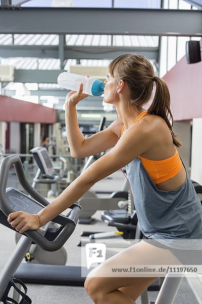 Young woman drinking water while exercising on a machine in gym