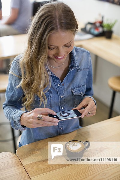 Glückliche Frau beim Fotografieren von Cappuccino mit Smartphone im Restaurant