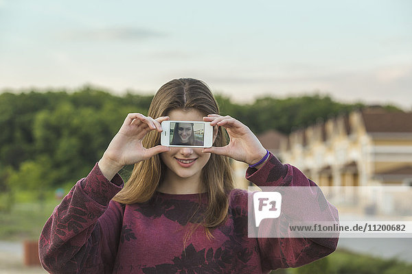 Fröhliche junge Frau zeigt Display von Smartphone mit ihrem Selfie im Freien