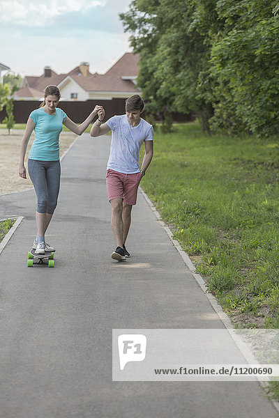 Mann assistiert Freundin beim Skateboardfahren auf dem Fußweg im Park