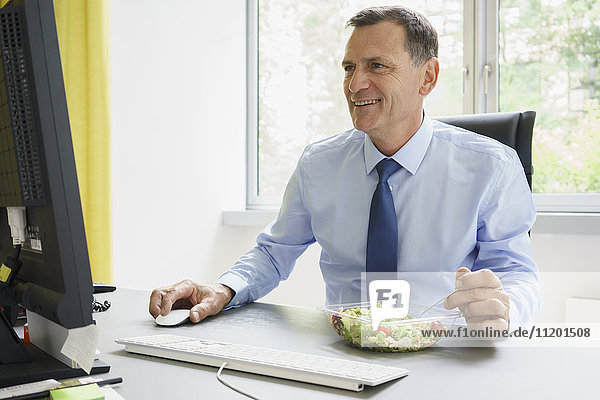 Lächelnder Geschäftsmann bei der Arbeit am Computer und Salat am Schreibtisch im Kreativbüro