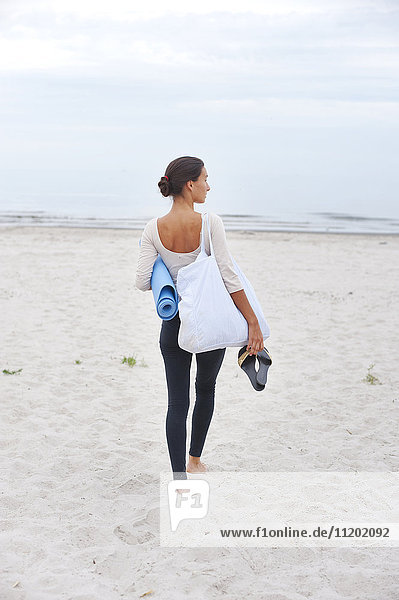 Young woman with exercise mat standing on beach