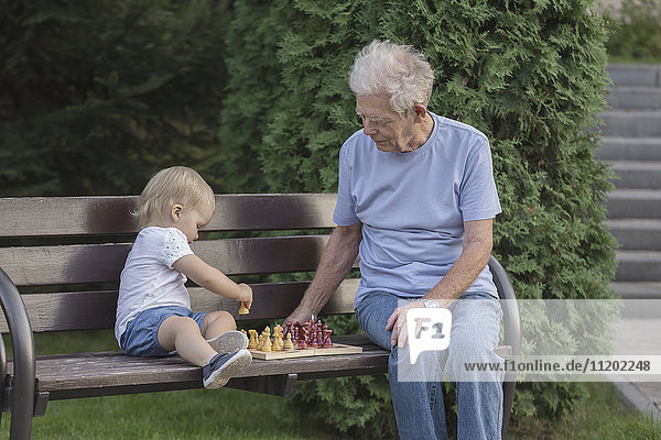 Senior Mann spielt Schach mit Enkelin auf Parkbank gegen Baum