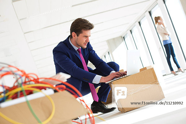 Young businessman crouching while using laptop on cardboard box in new office