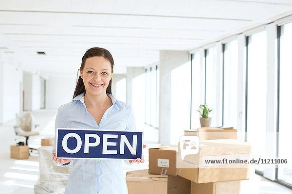 Porträt einer lächelnden Geschäftsfrau  die ein offenes Schild in einem neuen Büro hält