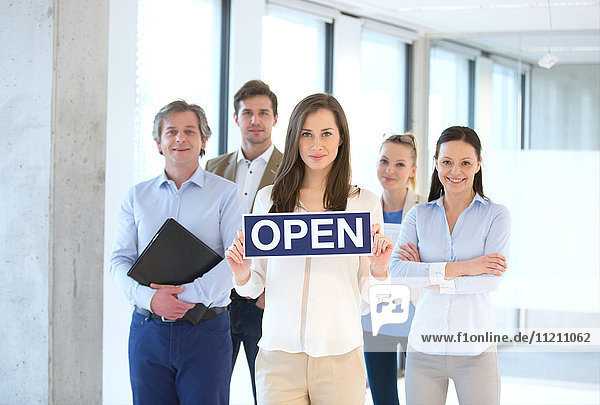 Porträt einer jungen Geschäftsfrau  die ein offenes Schild hält  mit einem Team im Hintergrund im Büro